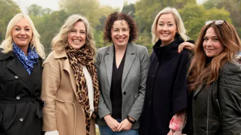 BBC/Graham Hodson Five women, wearing winter jackets and scarves, standing together in a park on an autumn day. All five are smiling at the camera.