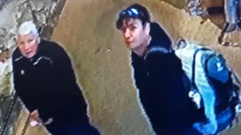 A blurry CCTV image taken from above shows two people stood side by side. They are both looking directly at the CCTV camera. One has white hair and wears all black. The other, who looks younger, has black hair and wears a black hoodie. He also has a light blue and dark blue rucksack on his back which appears to be full.