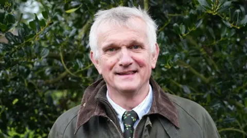 Qays Najm/BBC Nick Coller standing in front of a dense holly tree. He has short white hair and is wearing a green waxed jacket over a fleece and a white shirt with a black tie with pictures of holly on it