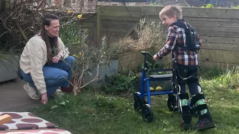 Asger, a young boy with blonde hair, is pushing a wheeled walking aid in front of him, while wearing an exoskeleton. The exoskeleton comprises leg braces and what looks like a power bank worn as a backpack. He is walking towards his mum Holly, who is crouching down to his level, in their garden.