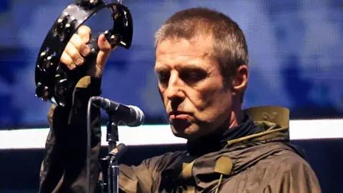 Liam Gallagher shaking a tambourine and looking impassively into the crowd on stage at the Cardiff Principality Stadium on 4 July 2025