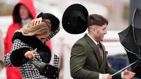 A woman in a black and white checked jacket holds her long blonde hair in place as her black hat flies off in windy conditions at the 2026 Cheltenham Festival. In front of her a young man in a dark green suit and with short brown hair smiles as the wind turns his black umbrella inside out