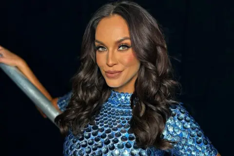 Mokkingbird Talent Vicky Pattison is slightly smiling, as if she's sharing an in-joke with someone, and is leaning against a scaffolding-style metal hand rail. Her right arm is resting up and long the rail. She's wearing a royal blue top or dress with a fish scale pattern and slightly shiny. She has long, dark, curly hair and full make up.