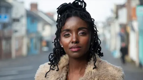 BBC News Young woman looks into the camera whilst standing on a town high street. She wears a coat with a furry collar and her black hair is half up and half down. 