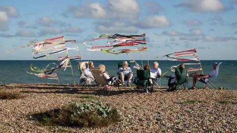 A group of retirees sitting on a pebble beach in deck chairs. There are flags and streamers next to them.