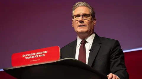 Sir Keir stood at a lecturn at Welsh Labour conference. 'Together for Wales' is written in England and Welsh on the lecture in front of him. He is wearing a black suite, red tie and white shirt.