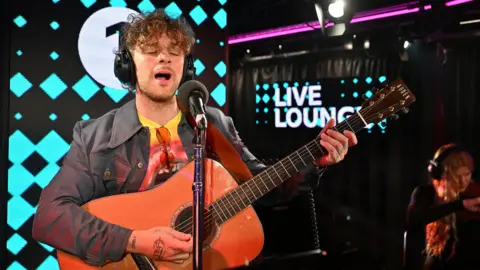 Tom Grennan is performing live in a studio, playing an acoustic guitar and singing into a microphone. He is wearing headphones and a denim jacket over a colorful yellow and pink t-shirt. Behind him a sign reads ‘LIVE LOUNGE’ with blue diamond-shapes on the wall. Another musician is in the background, also wearing headphones and playing an instrument.