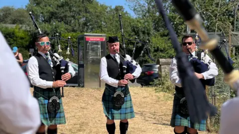 Three pipers wearing green and blue kilts, sporrans and dark waistcoats over white shirts. They are playing their pipes which are held on their left sides. Behind them is grass, parked cars, trees and blue sky.
