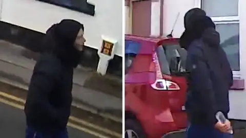 CCTV footage of a man wearing dark clothing. He has his hood up and is carrying a water bottle.