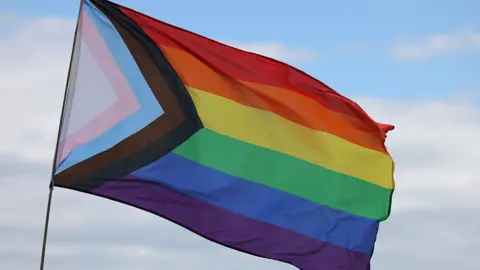 The Progress Pride Flag flying against a cloudy sky. It has the rainbow colours in stripes and a triangle at the side with a white triangle and stripes in pink, blue, brown and black