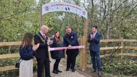 Shaun Pickering A man and a woman stand under a wooden archway in the middle of a wood, to cut a pink ribbon. Three other people next to them are clapping.