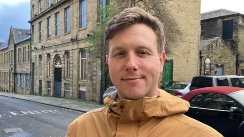 A man in a dull-yellow hooded top standing in a street with a row of buildings behind him