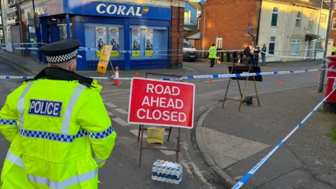 A police officer facing away from the camera looking at a cordon. There is a red road closed sign in front of him at a road junction which is also surrounded by police tape.