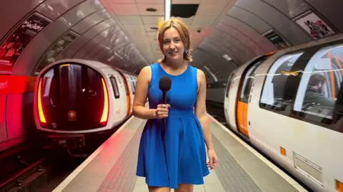 A girl with short ginger hair wearing a blue skater dress stands on the platform inside a Glasgow Subway station with two subways on either side.