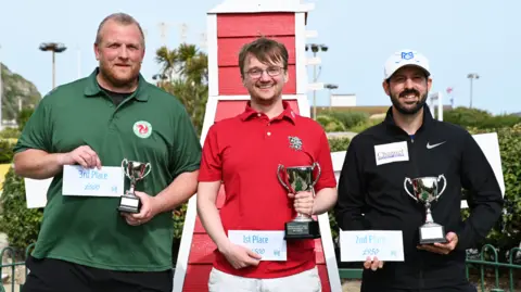 The image shows three men standing with trophies and certificates showing their podium placement. The winner, in the middle, is wearing a red shirt, the man in 2nd place on the right is wearing a black shirt and the last finalist on the left is wearing a green shirt. Behind them, bushes can be seen.