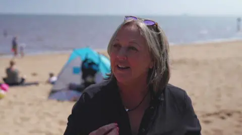 A woman with shoulder-length hair and sunglasses on her head. She is wearing a necklace and a dark-coloured blouse and is stood on a beach. A blue tent and people sitting on the sand can be seen behind her. The sea is in the distance.