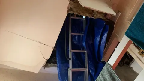 Hayley Johnson The ceiling and insulation of a house hanging down, with a ladder and blue sheeting in the gap left by the damage