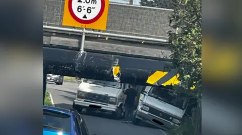 A low 2m (6ft 6in) high underpass, with circular red and white warning signs that has unseen railway track above.
The is a yellow and black chevron that is badly damaged by vehicles hitting it and two large camper van type vehicles are in the roadway beyond the tunnel and there is a blue car on the approach to the underpass in the middle of the road.