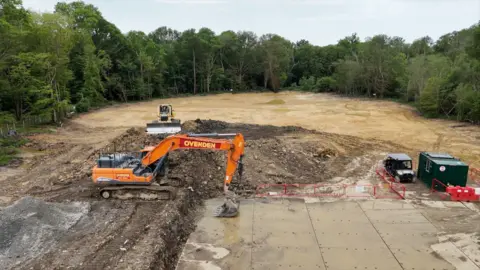 An aerial shot of a large swathe of brown muddy land, surrounded by green trees. In the foreground is an orange digger