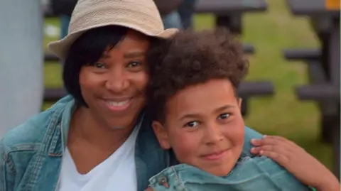 A mother and son. Hilary is sitting on the left and is looking directly at the camera and smiling. She is wearing a white T-shirt, denim jacket and beige hat. On her left is River, who is also looking directly at the camera and smiling. Hilary has her arm round him.
