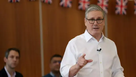 Reuters Keir Starmer has short grey hair and wears a white shirt. He points in the direction of the camera. Two men watch from behind him and union jack bunting is draped across a brown wall.