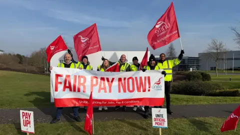 BMW workers on the picket line outside the BMW Hams Hall plant in Coleshill, holding up a large banner that says fair pay now. They also display red Unite the union flags.