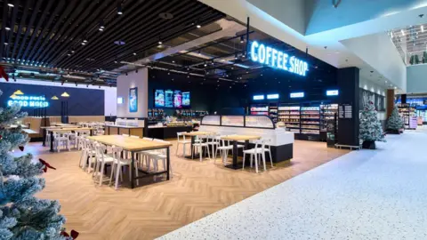 Marks and Spencer Image of a coffee shop, inside a department store. There are a number of wooden tables with white chairs, fridges of canned drinks and a food service area.