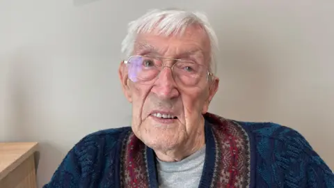 An elderly man with grey hair and glasses in a blue cardigan with dark red patterned trim