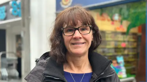 Adele, a woman with short brown wavy hair, she has a fringe and wears glasses.