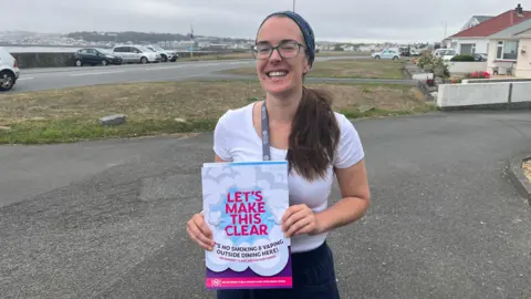 A woman with her hair in a ponytail and wearing a headband, holding a sign which says 'Let's Make This Clear'.