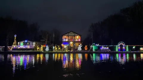 Robin Smith A home with multiple festive style light features, including snowmen, reindeers and trees, outside which are all glowing and reflecting on a body of water.