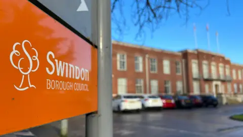 An orange Swindon Borough Council sign with a building behind it, with cars parked outside. The building in the background is out of focus.