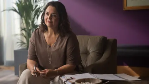 BBC | Mammoth Screen Actor Ruth Jones, seats in a chair with a file of papers on a desk and a pen in her right hand
