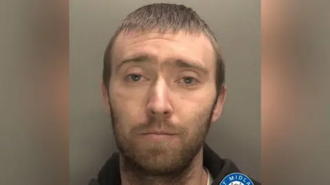 A mugshot of a man with short, brown hair and a beard. He is staring into the camera and wearing a black hoodie.