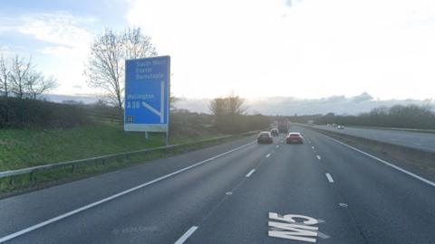 Google maps image of the M5 motorway in Somerset. A blue sign can be seen on the left indicating the exit for junction 26, Wellington. Some cars can be seen in the distance
