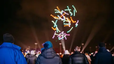 Yuup People are bundled up in jackets and woolly hats and are outside at night watching a drone display above them.