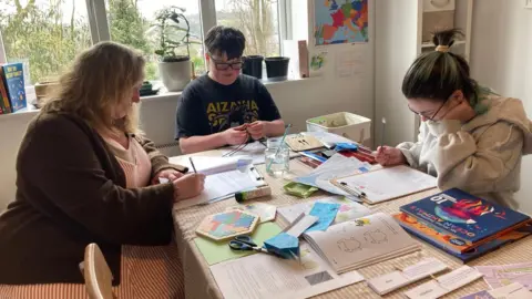 A middle-aged woman and two teenage girls sit working with pencils and paper at a table in a house. Educational material including books are on the table and trees can be seen outside through the window.