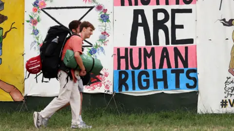Two men walk into Glastonbury Festival carrying a tent and sleeping bags. They are walking past a sign, which has the words 'are human rights' visible. 