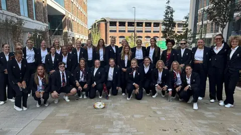BBC The full England women's rugby team are in two rows with those at the front kneeling down. Everyone is smiling at the camera and wearing matching jackets and white shirts.