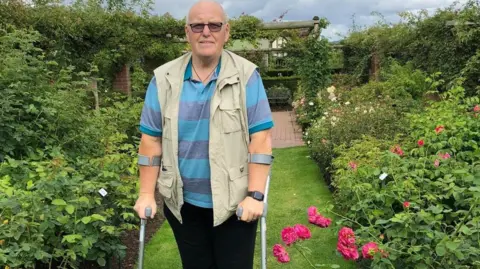 Cancer Research UK A man is using crutches in a garden. He is in the middle and bushes are to the left and right of him.