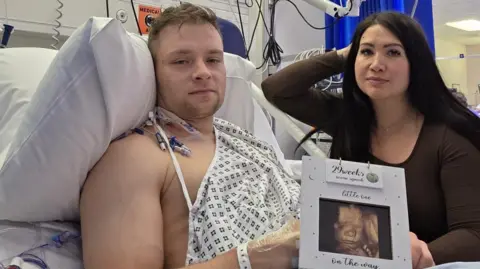 A man in a hospital gown lying on a hospital bed looks towards the camera holding a photograph of an ultrasound of his child, with a woman wearing a dark brown top and brown hair, by his side