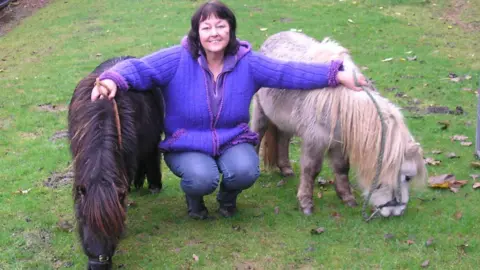 A dark-haired woman in a purple fleece and blue jeans, crouching down in field and resting her hands on two Shetland ponies, one black and one white.