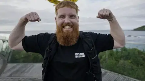 Russ Cook flexing his arm muscles as he stands in front of a yellow sign and a body of water. He is wearing a black t-shirt and looking directly at the camera.