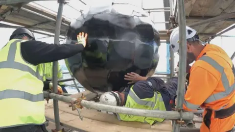 Workers wearing high visibility jackets and hard hats reinstall the black, shiny time ball on the Nelson Monument. 