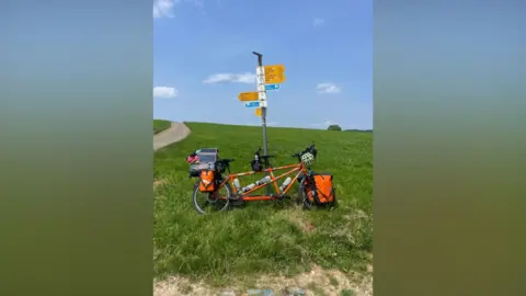 Chris and Nicky Clark An orange tandem, laden with luggage and propped against a fingerpost signing different places on yellow signs. It is in the middle of a large field, with only a narrow glimpse of a road to the left and a tree in the far distance