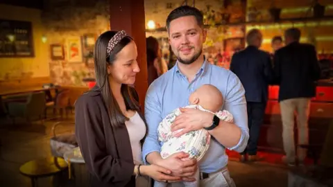 Ben Richmond A person in the foreground holds a baby dressed in a patterned onesie, with another person standing beside them and resting a hand on the baby. They are inside a spacious bar or restaurant with exposed brick walls, warm lighting, and a long bar counter in the background. Several people stand near the bar, and the room contains chairs, low tables, and decorative plants.
