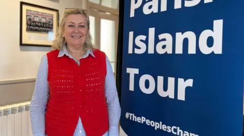 Deputy Helen Miles stands next to a large pop-up banner that reads Parish Island Tour #ThePeoplesChampion. She is smiling at the camera. She is wearing a pair of glasses on her head, a blue and white striped shirt and a red knitted waistcoat over it. She has shoulder length blonde hair. 