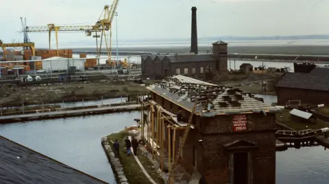 Canal & River Trust The Boat Museum at Ellesmere Port having its roof installed. In the distance is the River Mersey, a yellow crane and a pumphouse.