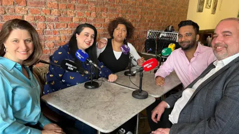 A group of people sit around a table. There are five people in the picture, smiling at the camera. Three women sit on one side and two men sit on the other. All have microphones.