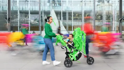 Sarah Walker Shot of a woman wearing headphones, a green top and jeans pushing a pram decorated with bright-green material with blurred coloured-shapes in the background